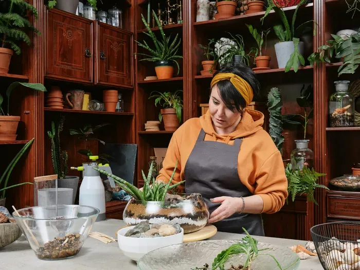 Woman florist in apron arranging green plants in glass bowl surrounded by pots and tools at her shop for Town Listing Hub